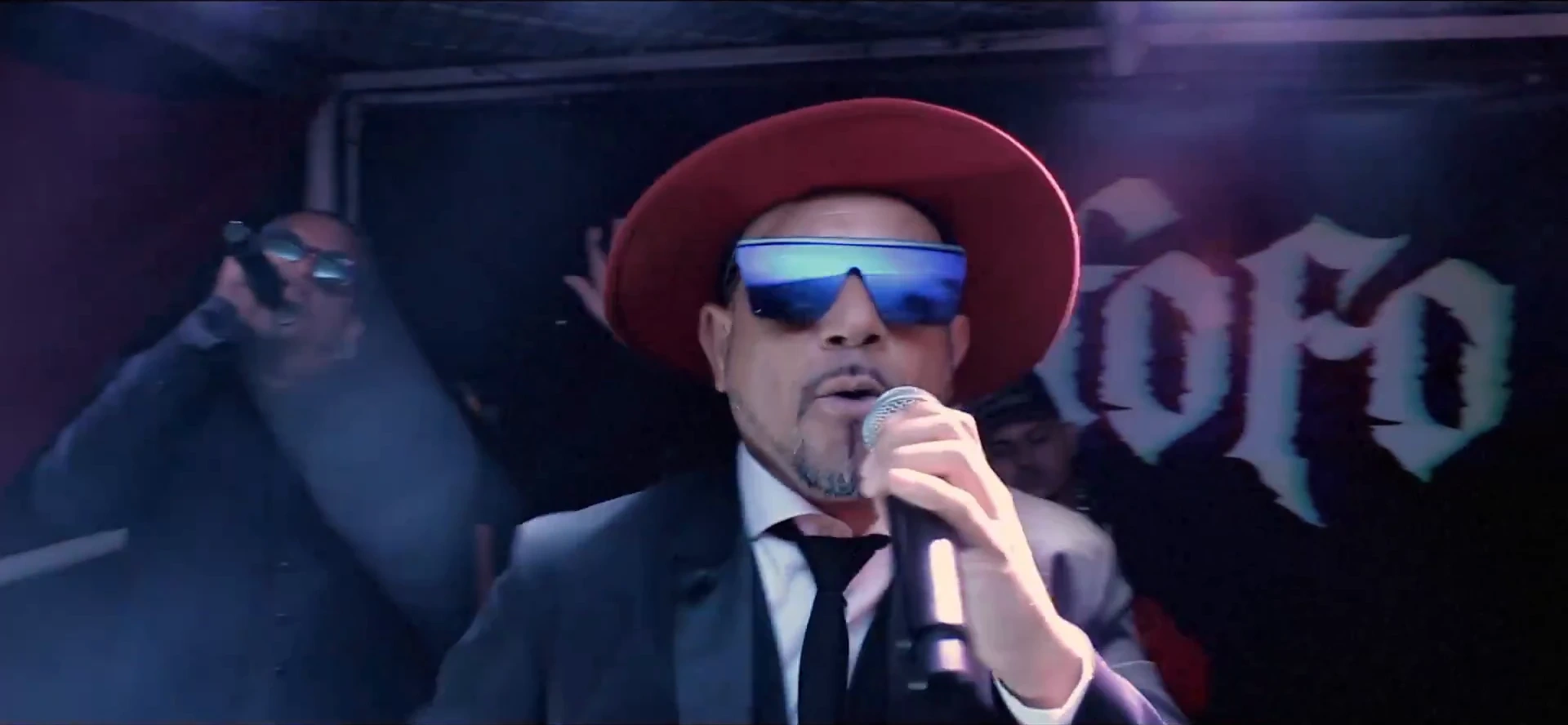 Close do cantor de chapéu vermelho e óculos espelhados azuis, cantando ao microfone, com o letreiro gótico "Cafofo" iluminado em violeta ao fundo. O vermelho do chapéu se destaca do azul-escuro do palco.