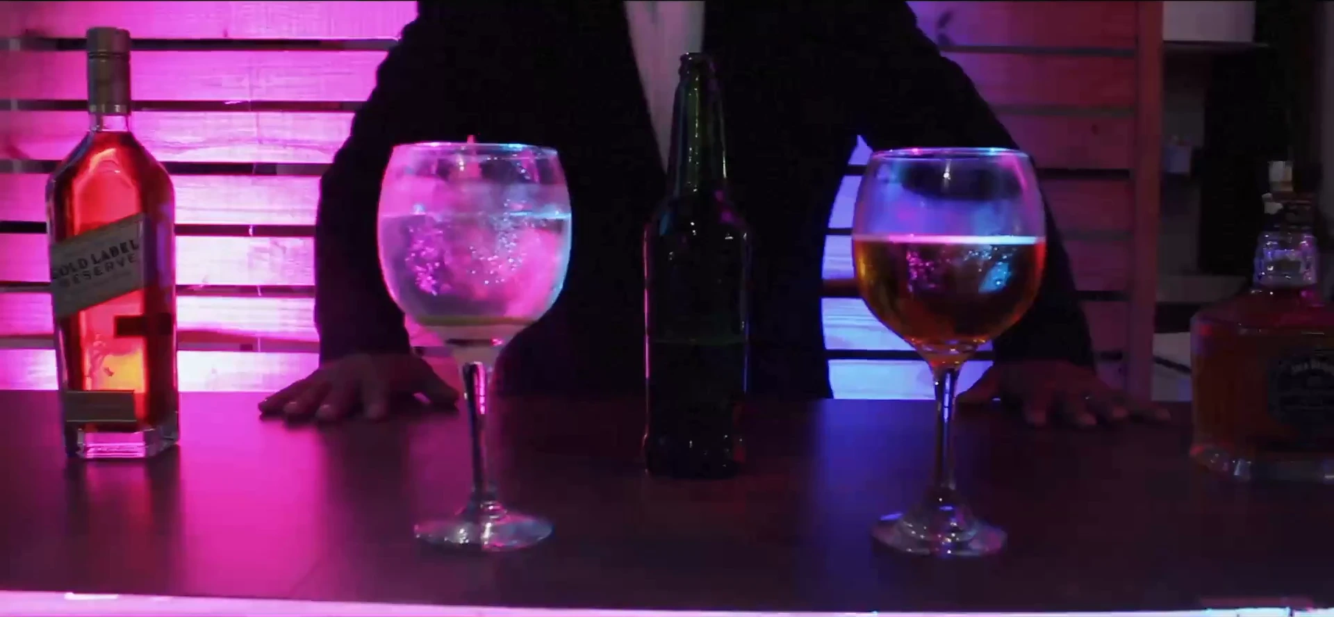 Balcão de bar iluminado em rosa e azul, com duas taças, uma garrafa e as mãos de uma pessoa apoiadas no tampo. O plano de detalhe quebra o ritmo do palco e da multidão.